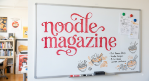 NoodleMagazine written in beautiful red lettering on a whiteboard