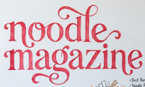 NoodleMagazine written in beautiful red lettering on a whiteboard